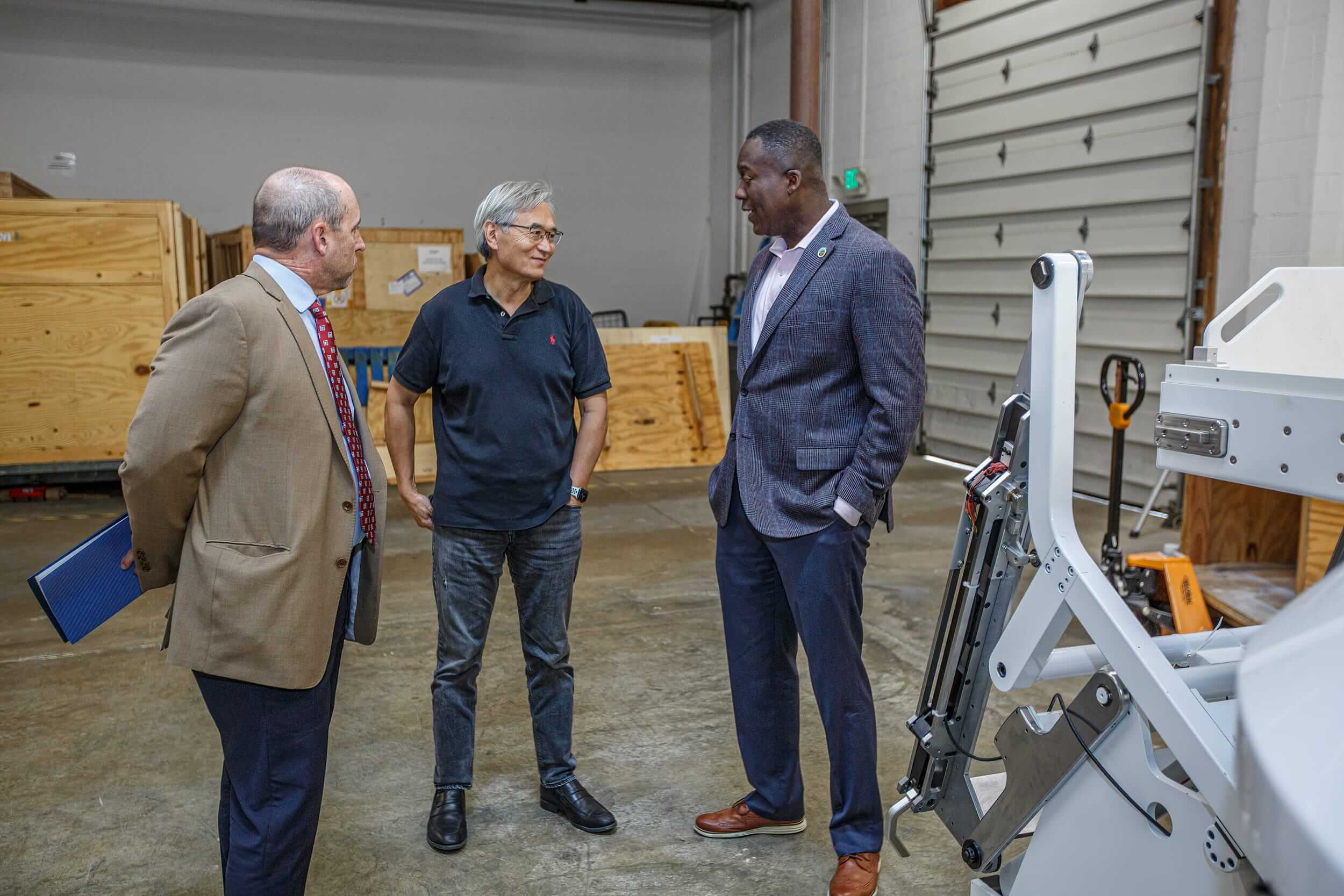 Two men in suits speak to another man in a black polo next to a piece of machinery in a warehouse.