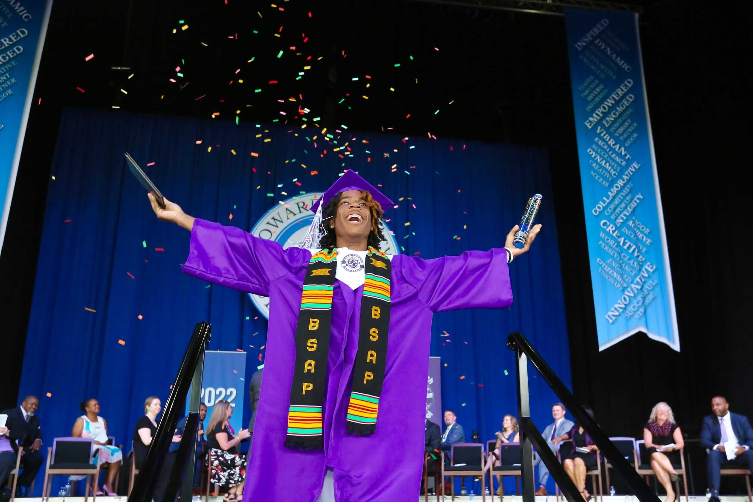 A student in a purple cap and gown smiles and celebrates while walking on stage with confetti going up behind him.