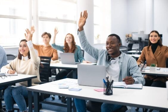 A group of students sitting at desks in a brightly lit classroom smile as they raise their hands to answer a question.