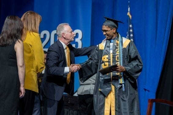A student in a black cap and gown shakes hands with an older man while receiving his diploma onstage.