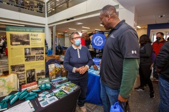 A tall man speaks to a masked older woman manning a table at a convention on sustainability.