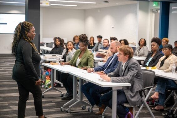 A suited woman engages with an audience of business people sitting in rows of desks.