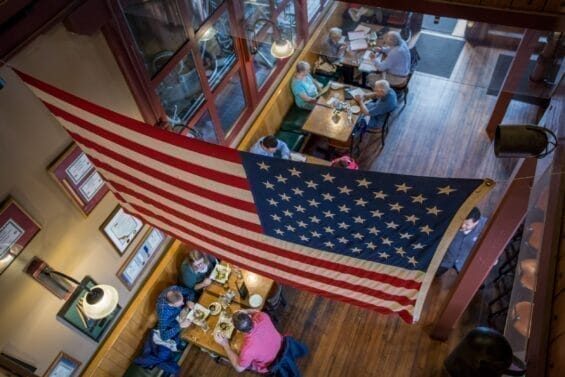 An American flag is visible in an aerial view of a brewery filled with people sitting together and enjoying food and drinks.