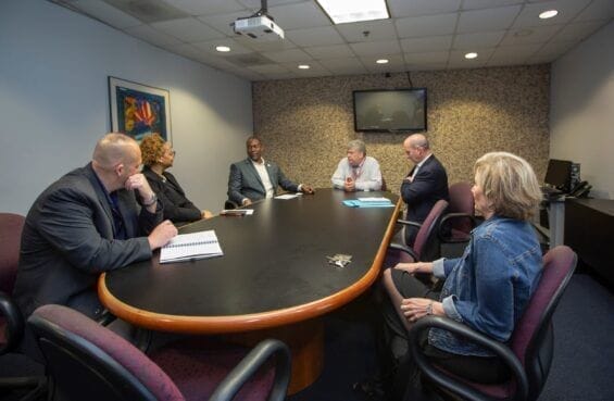 A diverse group of business people sit around an oval desk in a board room.