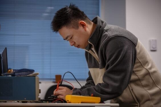 A man leans over a desk holding a red and a black wire to repair a piece of hardware.
