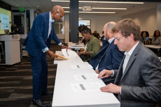 A suited man collects paper forms from a row of business people sitting at a long desk together.