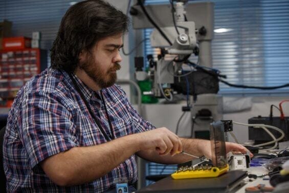 A bearded man sits at a desk and fine tunes a piece of hardware.