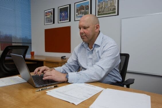 A business man sits at a desk and uses his laptop in an office room with a whiteboard behind him.