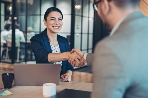 A woman smiles as she shakes hands with a person behind the camera.